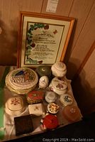 Photo of thirteen keepsake boxes including wooden, ceramic, and metal types with floral and figurative decorations, plus framed art with an inspirational text and vine border