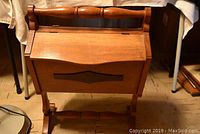 Front view of the maple knitting cabinet showing the hinged front panel and top lid, standing on turned legs with cross supports.