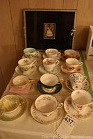 Twelve assorted vintage tea cups and saucers arranged on a table with a black tray standing upright in the background.