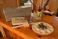 Photo showing clustered art supplies on a wooden table, including a plastic storage box, sets of oil pastels, paint brushes in a holder, and a ceramic mixing palette.