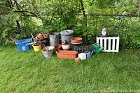 Photo shows entire collection of gardening items arranged on grass near shrubbery including metal garbage cans, plastic pots and planters, wooden baskets, driftwood, and garden edging.