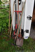 Photo showing assorted garden tools leaning against white siding including shovels and rakes.