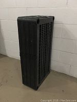 Collapsed black plastic shelving unit placed upright next to a beige brick wall showing its grid structure and compact storage form.