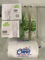 Photo showing unopened boxes of 500 plastic forks and knives labeled Café Express, two packs of 50 biodegradable plastic cups, and a large pack of Royal Chinet paper plates.