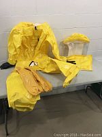Yellow hazmat suit laid out on a table with hood and gloves visible. Shows full suit, hood with clear face shield, and two pairs of gloves.