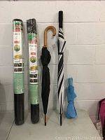 Photo shows three umbrellas of varying types and two unopened rolls of Jobe's landscape fabric stacked vertically against a wall.