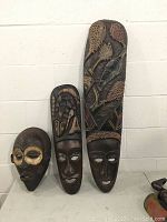 Three carved wooden masks arranged on a surface against a white wall: one round mask and two long masks, displaying varying ethnic designs and carvings.