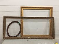 Photo of three retro picture frames leaning against a wall: one large gold, one large taupe, and one small oval brown frame.