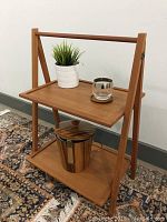 Side view photo of the teak folding side table with two shelves, holding a small potted plant, glass cup, and metal pitcher as display items.