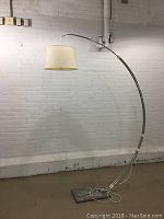 Full view of vintage large arc floor lamp with curved metal arm and cream fabric lampshade over a metal base on concrete floor against brick wall.