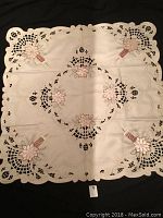 Top view of square white cutout tablecloth with pink embroidered floral and candle motifs and gold trim