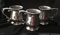 Three pewter mugs with ornate handles shown together on a black fabric background to display shape and condition.