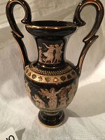 Front view of black Greek-style vase on white cloth with detailed painted ancient Greek figures and gold trims on handles and rim.
