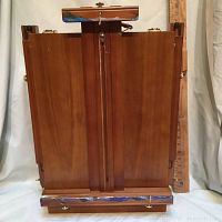Closed travel art easel in vertical position showing polished wood front with leather handle on top and adjustable screw mechanisms.