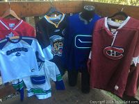 Display of multiple hockey jerseys on hangers including youth and adult sizes with logos from Canada, Canucks, Montreal Canadiens, Boston Bruins