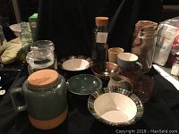 View of assorted kitchen pottery items including plates, bowls, mugs, and glassware in earth tone colors arranged on black cloth.