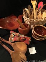 Photo showing large burl wood bowl, wooden mortar and pestle, small ceramic bowls, giraffe carved wooden salad forks, fabric napkins with wooden rings and basket of wooden utensils on black surface.