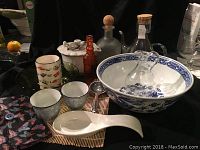 Lot showing large blue and white Oriental bowl with two glass pour bottles inside, white ceramic spoon in front, porcelain cups, multicolored fish cup, green glass plates, white lidded container with tea strainer, and amber glass bottle in back