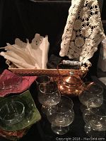 Full view of arranged lot on black cloth background showing copper teapot, straw basket holding napkins, four brandy glasses, and crochet tablecloth draped. Red and green square plates also partially visible.