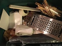 Top-down view of one Rubbermaid container showing assorted clothespins, rolls of white fabric or tape, a metal vent cover with grille design, and other hardware items packed inside.