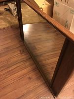 Front view of large rectangular mirror with wood frame showing reflective glass and wood edges, resting on a hardwood floor.