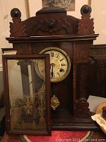 Front view of vintage wooden clock with open glass door showing etched decorative design and aged Roman numeral clock face with hanging pendulum inside.