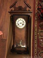 Front view of the vintage wall clock showing its long rectangular wooden case with a glass door, the clock face with Roman numerals, and decorative finials at the top.