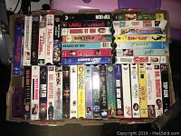 Photo showing assorted VHS movies stacked horizontally and vertically in a cardboard box including Top Gun, Mr. Bean, Stand By Me, and various other titles.