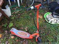 Red Razor youth electric scooter on grass. View shows handlebar, deck, and part of rear wheel, revealing rust and outdoor wear.