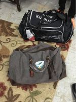 Two duffel bags side by side on carpet floor showing the black OGIO duffel bag and gray Miami Dolphins crossbody duffel bag