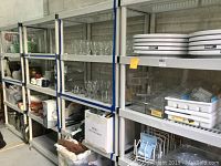 Four tall grey plastic shelving units placed against a concrete wall, holding various glassware and household items, demonstrating shelving structure and size.