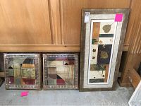 Three framed pictures: two square abstract geometric and one rectangular leaf design, set against wood panel background on floor.