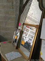 Three assorted mirrors leaning against a cinder block wall in a storage area. Large mirror has a dark stained wood frame with an ornate arched top and etching on the mirror surface. Medium and small mirrors have simple gold frames.