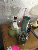 Five ceramic vases shown on table with different colors and shapes including green vase with handles, tall blue vase, and pink vase.