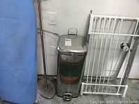 Photo showing blue ironing board, silver step garbage can, white towel rack in corner