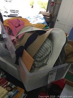 Plastic container filled with folded scarves, belts, and hats including a large white and brown hat among other fabric items.