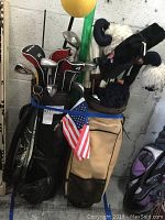 View of two golf bags side by side standing upright. Black golf bag on left side with several golf clubs and some headcovers. Tan golf bag on right side with clubs and plush animal headcovers. Small American flag attached to tan bag.