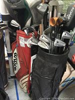Two golf bags with various golf clubs inside, including irons and drivers. One bag is black with an Adams driver cover, and the other is a red and white Wilson bag.