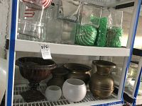 Two shelves with assorted brass urns and glass vases. Bottom shelf has brass urns and white glass vases; top shelf has clear glass containers including some filled with green decorative material.