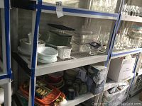 Shelf containing clear glass serving platters, green glass plates, white ceramic dishes, mugs, and Corning Ware items.