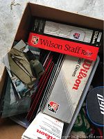 Box containing stacked white and black Wilson Staff Soft golf gloves in original packaging, with some red and black packaging visible, showing quantity and brand