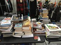 Wide view of multiple stacks of books on a table, showing the quantity and variety of books in the assortment including cookbooks and other genres