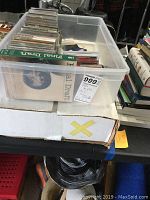Clear plastic storage bin holding a mix of media including VHS tapes, CDs, and possibly mini cassettes, labeled with an auction lot tag.