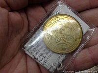 Close-up photo of the gold-colored $5 Kingston coin sealed in a plastic container held in hand, showing the building and fireworks design with text.