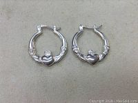 Pair of silver hoop earrings with Claddagh design on beige background, showing details and condition