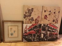 Photo showing one framed embossed botanical art piece with gold frame and double mat, plus four square canvas prints grouped beside it: two floral botanical and two street cafe scenes with red umbrellas.