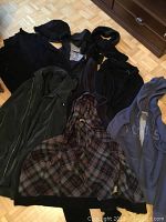 Six clothing pieces laid on floor including three coats, two sweaters, and a black knit hat. Visible plaid patterned sweater and gray Roots hoodie.