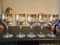 Five clear stemware wine glasses with gold-colored decorative trim on the rim shown upright in a row.