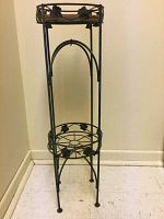 Full view of the tall metal stand with decorative leaves and stone top, standing against a wall on a tiled floor.