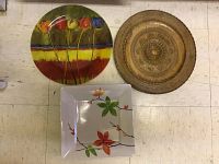 Three decorative platters/plates including one round floral-painted ceramic plate, one round metallic ornate tray, and one square white ceramic plate with floral prints.